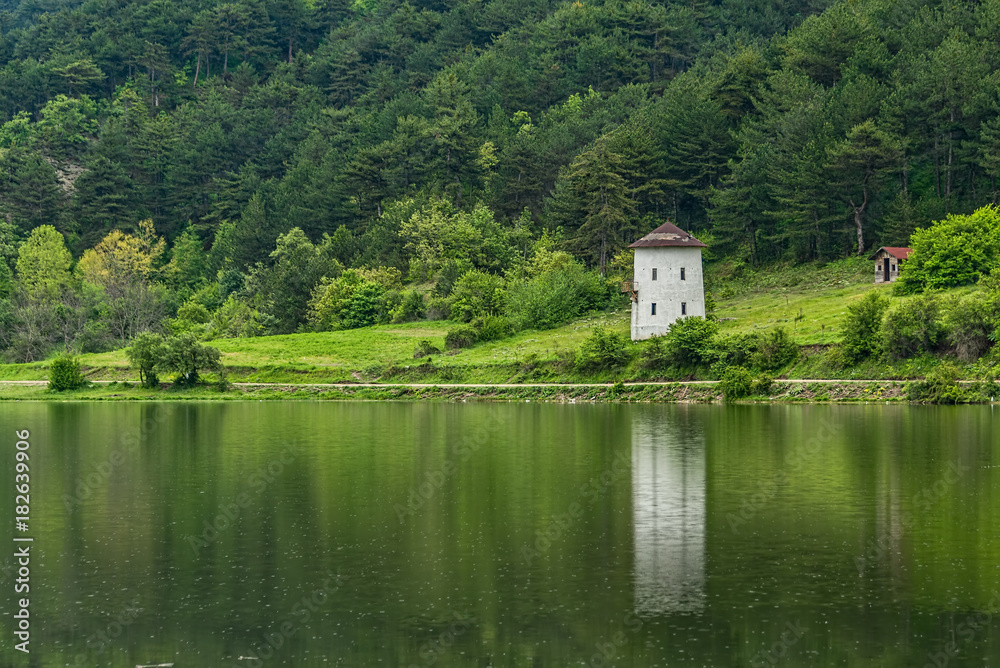 Fototapeta premium Reflections of green trees and an old windmill into a calm lake