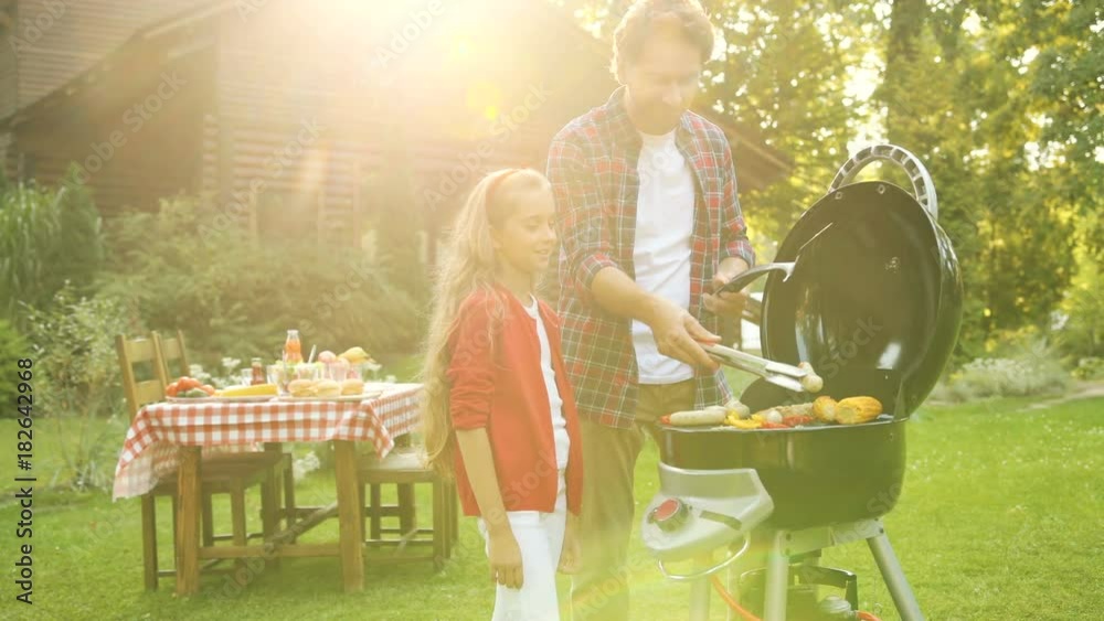 Handsome man grilling on the barbecue and his daughter watching him. Beautiful suuny day and nice hoyse on the backgrond. Outdoor
