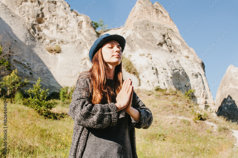 Beautiful young woman in hat and gray sweater meditates with folded hands and closing eyes against backdrop of beautiful mountains and hills on sunny day