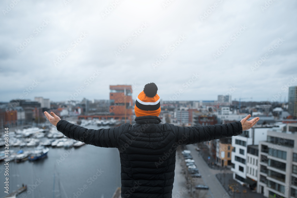 A young guy look at city from the roof of building with his arms raised. Black jacket and orange cap. Belgium, Antwerp.
