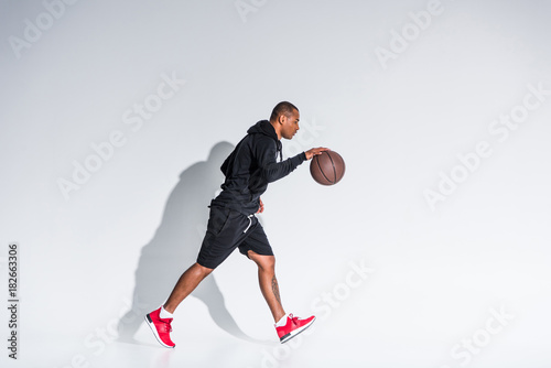 side view of young african american sportsman playing with basketball ball on grey