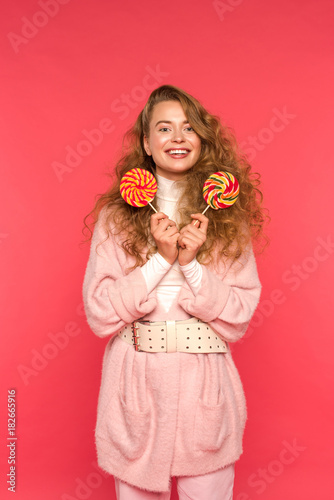 happy stylish girl standing with two lollipops and looking at camera isolated on red