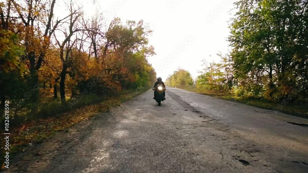 Unrecognizable man in black helmet and leather jacket approaching while riding a motorcycle on a road on a sunny day in autumn. Then he outruns the camera point of view and moves forward Unrecognizable man in black helmet and leather jacket approaching while riding a motorcycle on a road on a sunny day in autumn. Then he outruns the camera point of view and moves forward