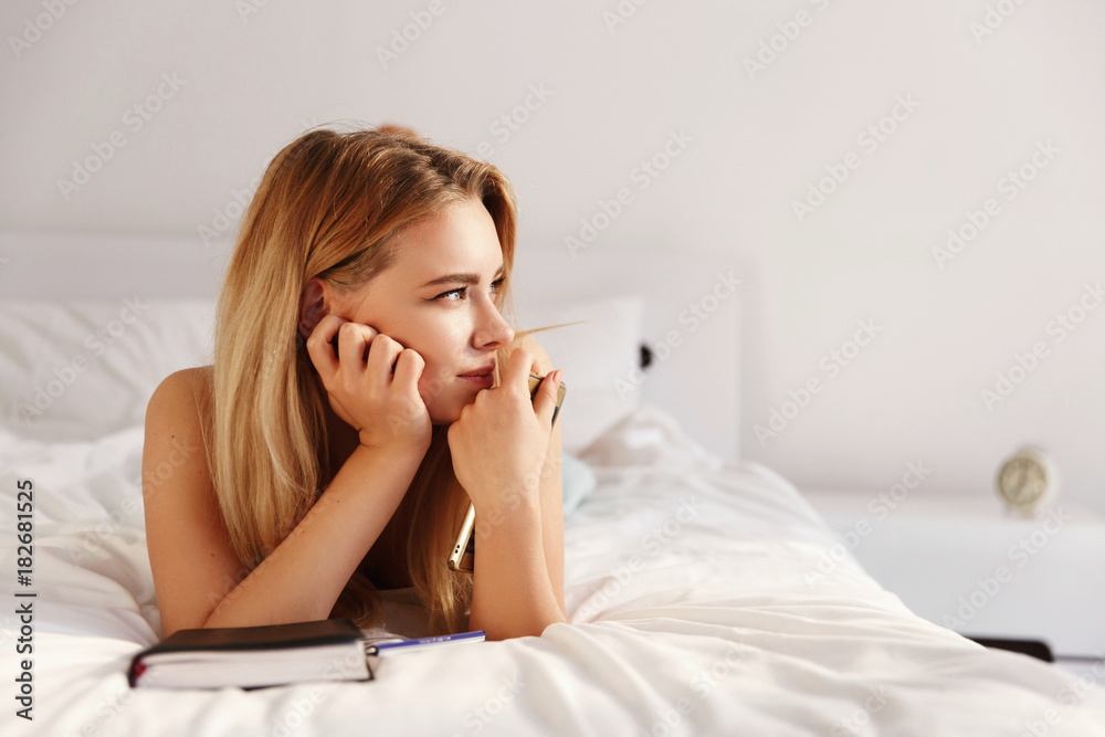 Beautiful blonde woman looks thoughtful sitting on the white bed with a notebook in the morning Beautiful blonde woman looks thoughtful sitting on the white bed with a notebook in the morning
