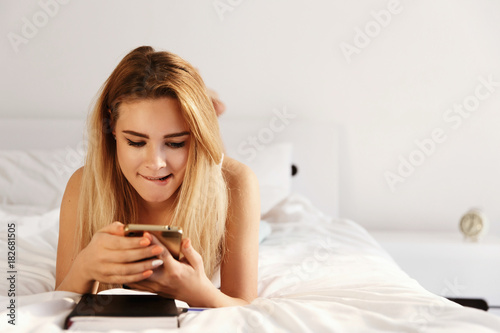 Beautiful blonde woman looks happy  lying on the white bed with a notebook in the morning and checking her phone
