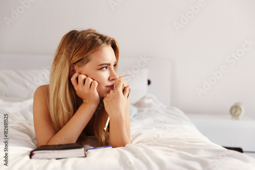 Beautiful blonde woman looks thoughtful sitting on the white bed with a notebook in the morning