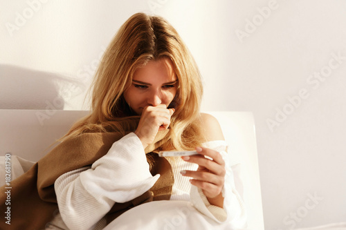 Ill blonde woman lies with a scarf in the bed and checks her temperature