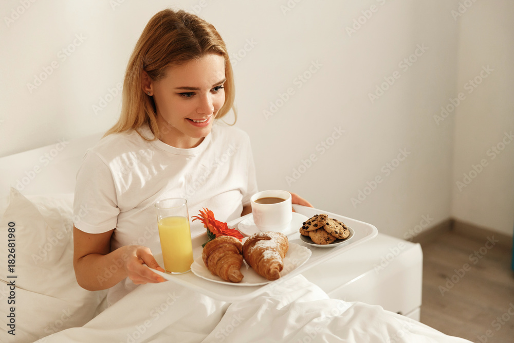 Blonde woman lies in the bed holds a tray with delicious french breakfast