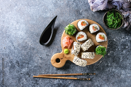 Homemade sushi rolls set with salmon, sesame seeds serving on wood serving board with pink pickled ginger, soy sauce, wasabi, seaweed salad, chopsticks on gray texture background. Top view, space