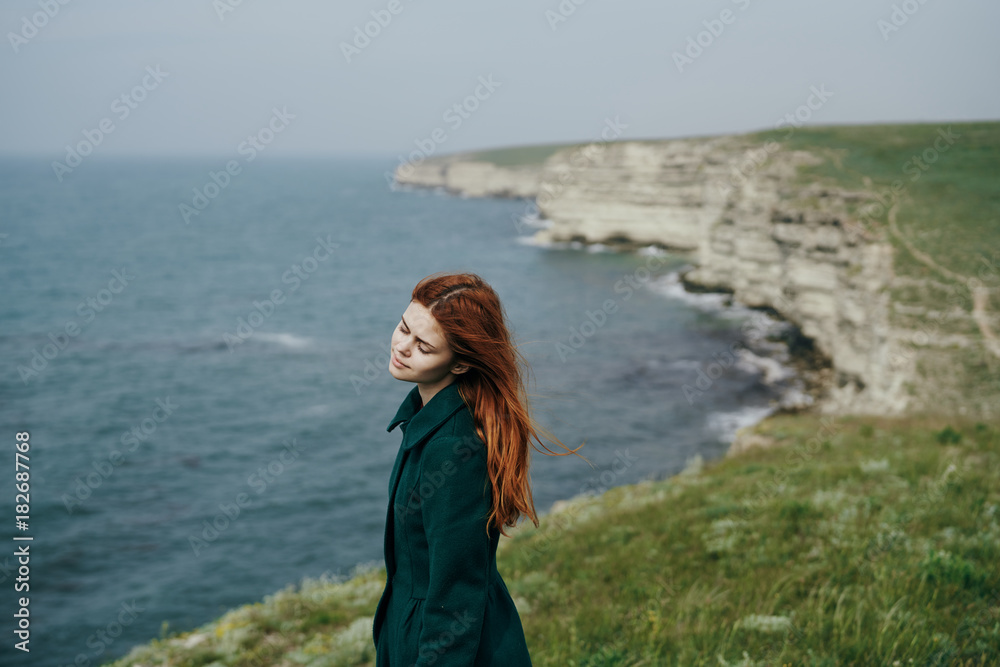 beautiful red-haired woman in a green coat is standing on a cliff, grass, sea, nature
