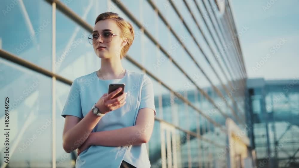 Stylish young woman in trendy sunglasses uses her mobile phone, outside the glassy urban building on a bright sunshine. Blogger, modern lifestyle Stylish young woman in trendy sunglasses uses her mobile phone, outside the glassy urban building on a bright sunshine. Blogger, modern lifestyle