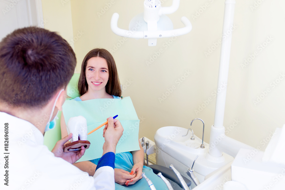 Young dentist male man with 3d model of one white big teeth explaining teeth treatment for young smiling female woman patient in dentist workplace in clinic Young dentist male man with 3d model of one white big teeth explaining teeth treatment for young smiling female woman patient in dentist workplace in clinic