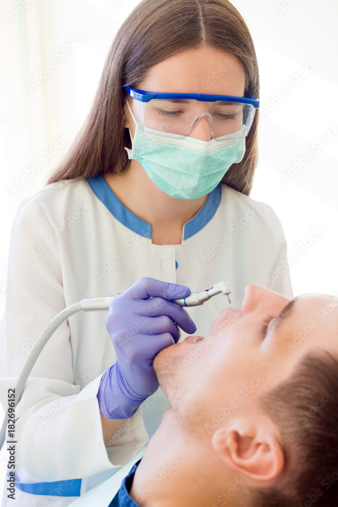 Young woman female dentist in medical glasses and mask with dentist equipment tool machine preparing for treatments teeth of smiling man male patient Young woman female dentist in medical glasses and mask with dentist equipment tool machine preparing for treatments teeth of smiling man male patient