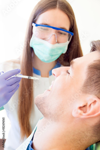 Young woman female dentist in medical glasses and mask with syringe of Anastasia preparing for treatments teeth of man male smiling patient