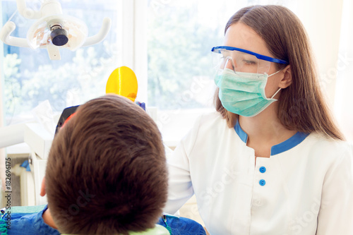 Young woman female dentist in medical glasses and mask treatments teeth of man male patient in red uv glasses by uv equipment tool implanting medical sealants