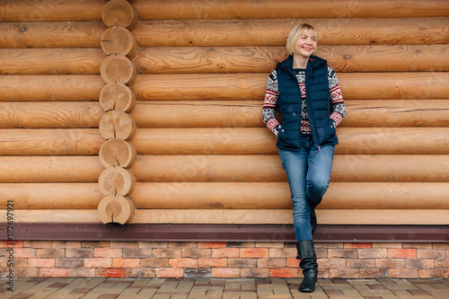 Cute pretty blonde woman in winter sweater and jeans standing near log house. Cheerful girl in vest. Wooden background