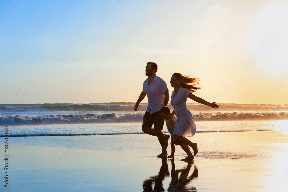 Fototapeta premium Happy full family - father, mother, baby son have fun together, child run with splashes by water pool along sunset sea surf on black sand beach. Travel lifestyle, parents with kids on summer vacation.
