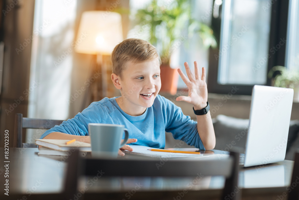 Distance communication. Happy excited young boy smiling and waving his hand while greeting his friend