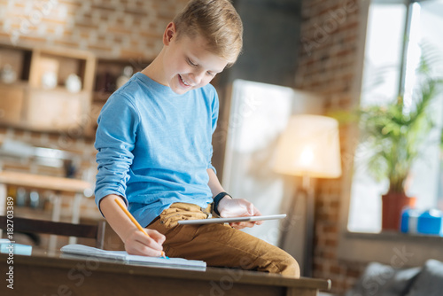 Taking notes. Delighted nice positive boy holding a tablet and taking notes while sitting on the table