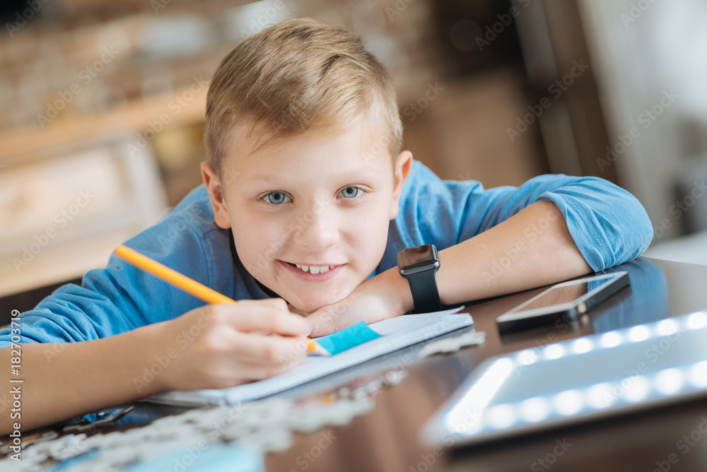 Interesting studying. Cheerful nice pleasant boy leaning on the table and writing with his pencil while studying at home Interesting studying. Cheerful nice pleasant boy leaning on the table and writing with his pencil while studying at home
