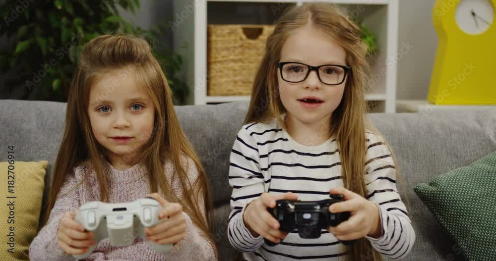 Portrait shot of cute small girls playing video games and holding video controls on the nice couch in the modern living room. Indoor Portrait shot of cute small girls playing video games and holding video controls on the nice couch in the modern living room. Indoor