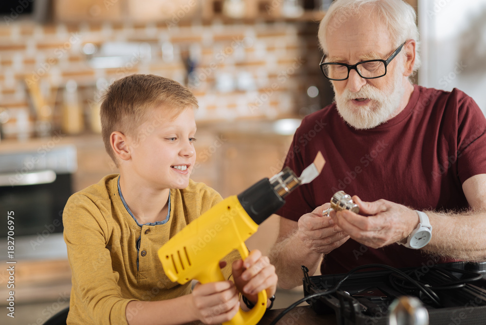 Professional skills. Nice pleasant senior man sitting together with his grandson and holding a drill detail while teaching his grandson to use it Professional skills. Nice pleasant senior man sitting together with his grandson and holding a drill detail while teaching his grandson to use it
