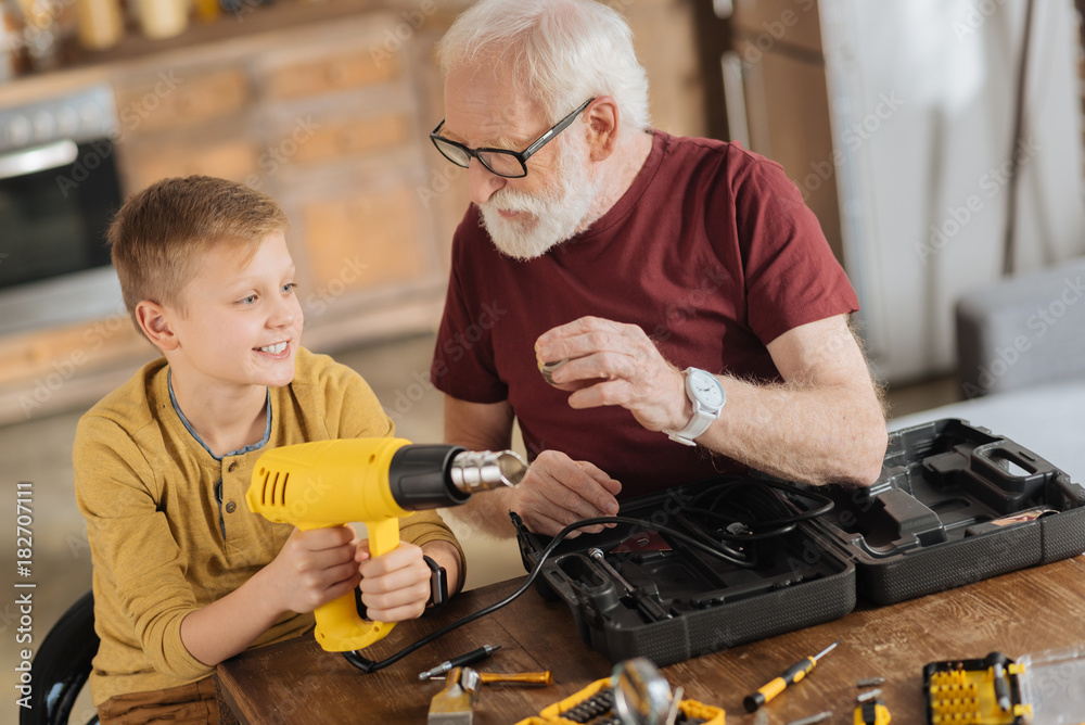 Male skills. Joyful nice positive boy sitting next to his grandfather and holding a drill while helping him