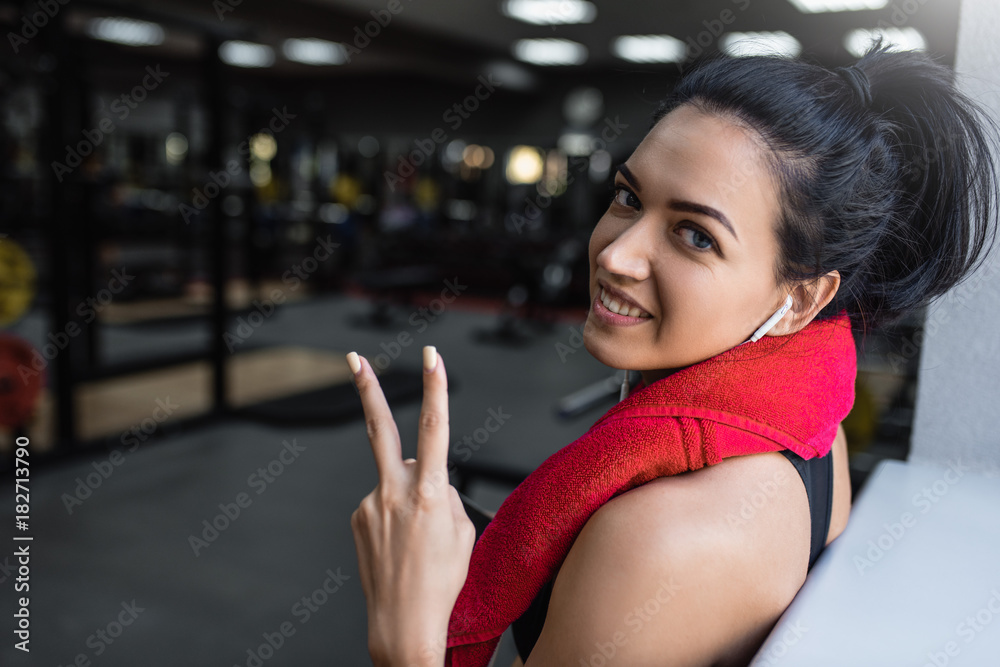 Beautiful shot of happy young beautful fitness woman taking a break from her workout with red towel on neck and earphones listening the music and showing peace sign in the gym.