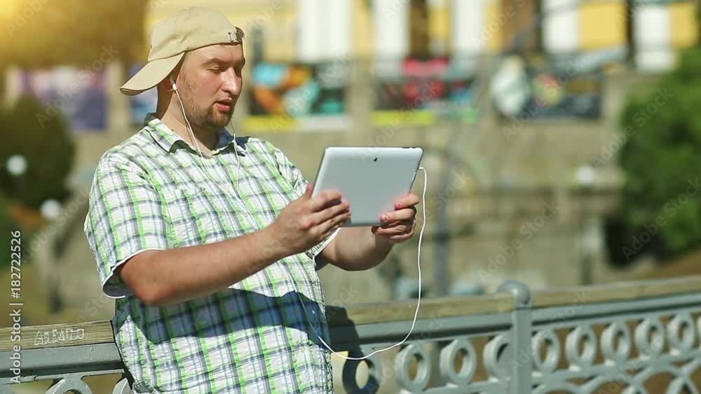 Man communicates via tablet computer. Fat boy in checkered shirt and baseball cap holds in hands white tablet pc with earphones. Man with tablet pc communicates through videochat Man communicates via tablet computer. Fat boy in checkered shirt and baseball cap holds in hands white tablet pc with earphones. Man with tablet pc communicates through videochat