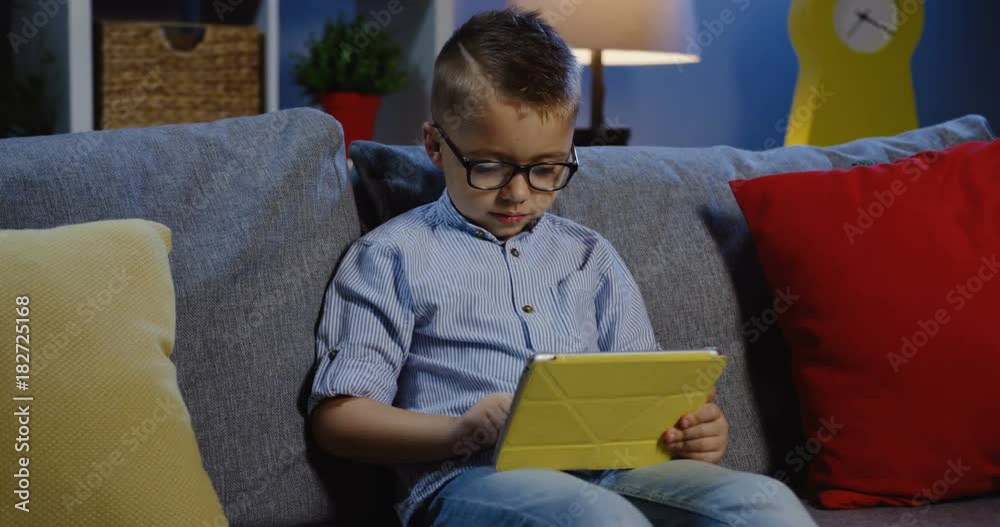 Portrait shot of the small blonde boy in glasses sitting on the grey sofa with pillows in the nice room at home and typing on his tablet device. Inside Portrait shot of the small blonde boy in glasses sitting on the grey sofa with pillows in the nice room at home and typing on his tablet device. Inside