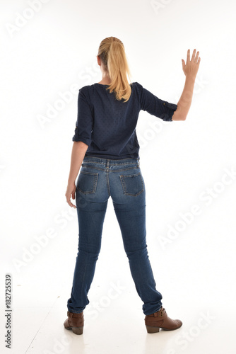 Full length portrait of a girl wearing simple blue shirt and jeans, standing pose facing away on a white background.