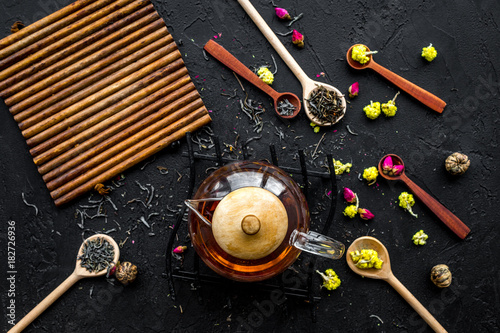Brew the aromatic tea. Tea pot near wooden spoons with dried tea leaves, flowers and spices on black wooden background top view mockup