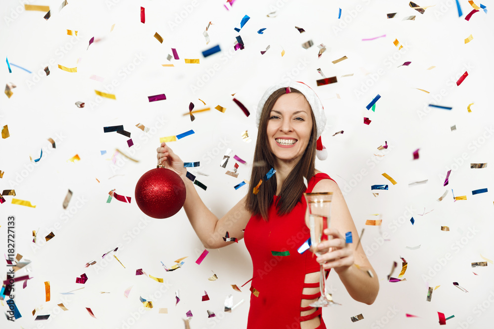 Fototapeta premium Beautiful smiling caucasian young happy woman in red dress and Christmas hat standing with big tree toy and glass of champagne on white background confetti. Santa girl isolated. New Year 2018 holiday