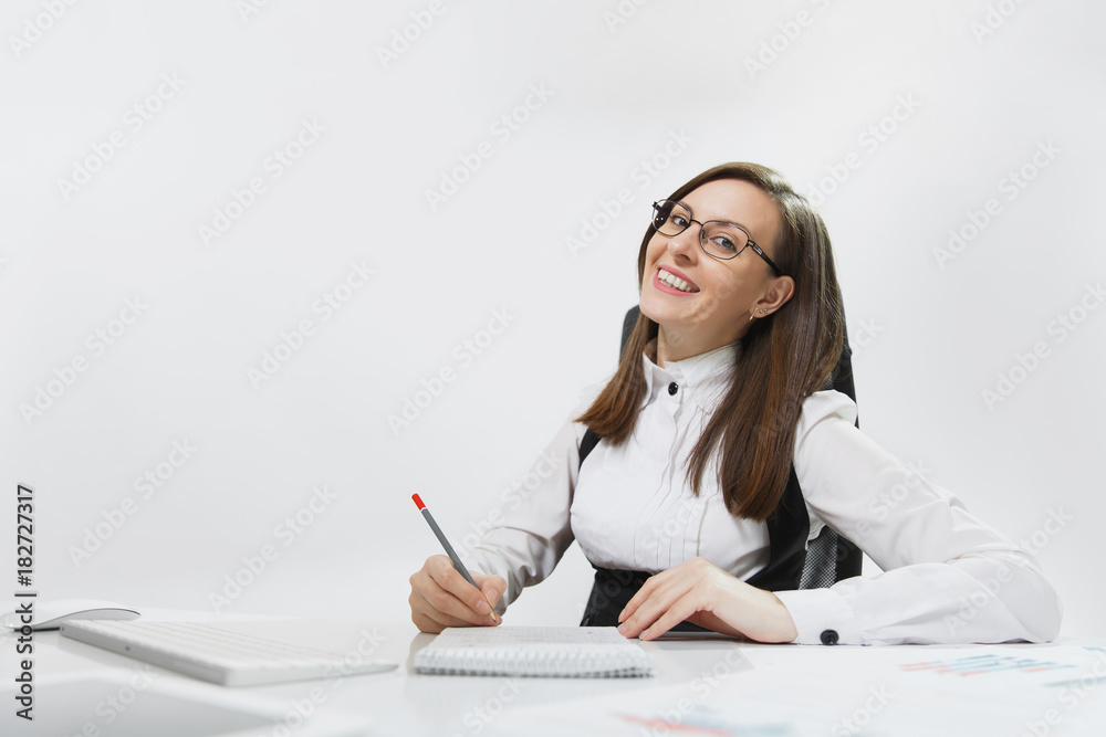 Beautiful smiling brown-hair business woman in suit and glasses sitting at the desk, working at computer with modern monitor and documents in light office, writing in notebook on white background Beautiful smiling brown-hair business woman in suit and glasses sitting at the desk, working at computer with modern monitor and documents in light office, writing in notebook on white background
