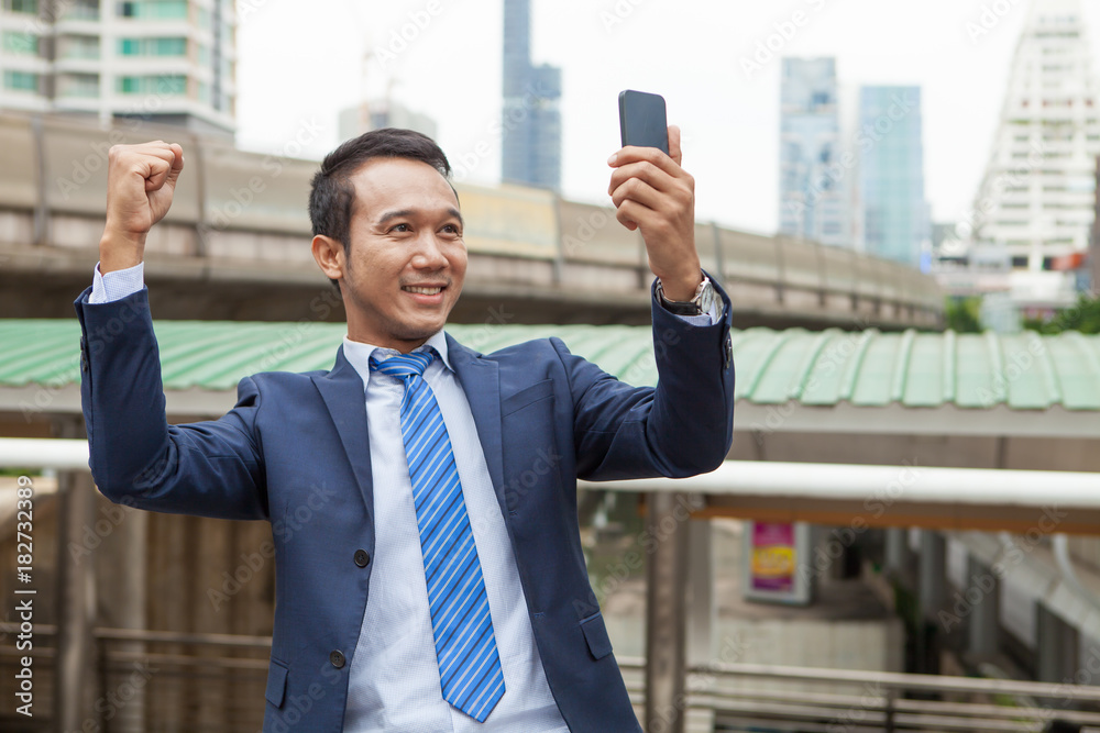 Businessman raising his fist in the air while looking on mobile phone, with office building background - business success, achievement, and win concepts Businessman raising his fist in the air while looking on mobile phone, with office building background - business success, achievement, and win concepts
