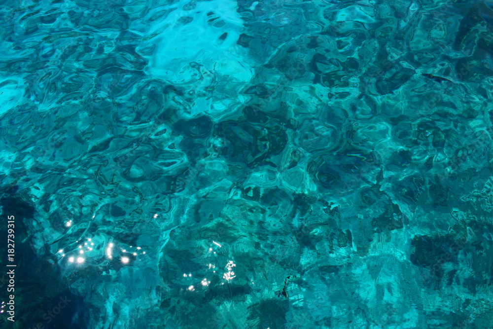 Background of transparent azure sea water with sunlight reflecting on a summer day. On the surface of the water a soft wave pattern.