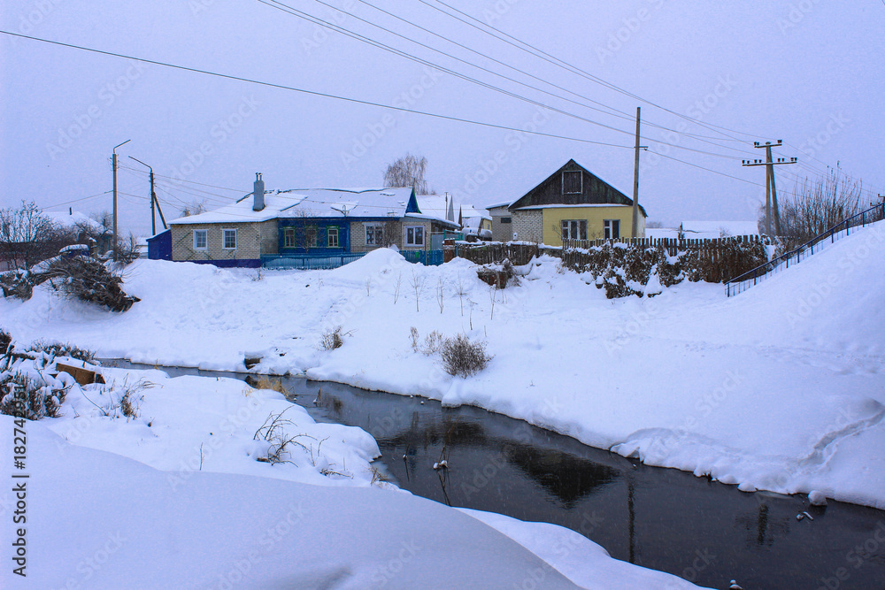 The river is not yet completely frozen and flows on the outskirts of a snow-covered Russian village in the winter twilight