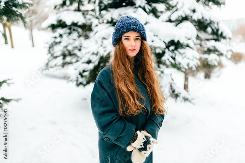 Young beautiful teenager girl in oversized wool coat with long brown beautiful hair standing in winter city park with snowy spruces on background. Fashionable stylish lady with amazaing blue eyes.