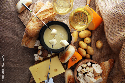 Gourmet Swiss fondue dinner on a winter evening with assorted cheeses on a board alongside a heated pot of cheese fondue with two forks dipping bread and white wine behind in a tavern or restaurant