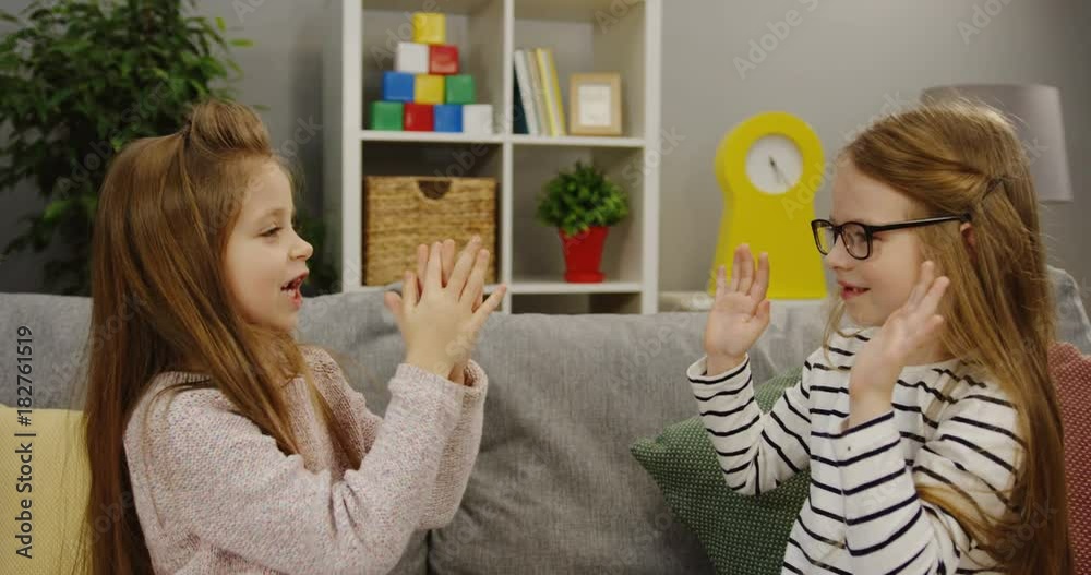 Two cute similar girls playing hands game on the couch in the living room. Friends having fun at home. Indoor Two cute similar girls playing hands game on the couch in the living room. Friends having fun at home. Indoor
