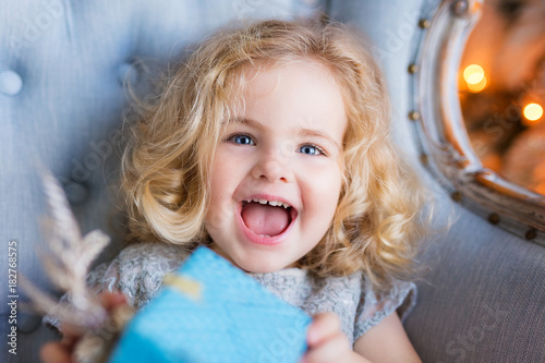 Beautiful happy little girl smiling and holding a present box. Сlose-up portrait. Christmas. New Year. Indoor. Home. 