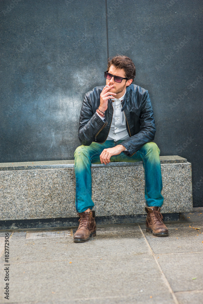 Wearing black leather jacket, blue jeans, brown boot shoes, sunglasses, a  young guy with beard sitting on marble bench in corner, smoking cigarette  during working break, trying mind calming down. Stock Photo