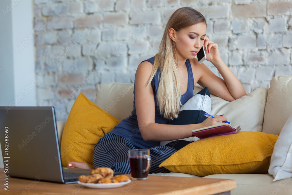 Female freelancer in her casual home clothing working remotly from her dining table in the morning. Houses on a sofa on a white brick wall background. Architect, blueprints. talking on the phone Female freelancer in her casual home clothing working remotly from her dining table in the morning. Houses on a sofa on a white brick wall background. Architect, blueprints. talking on the phone