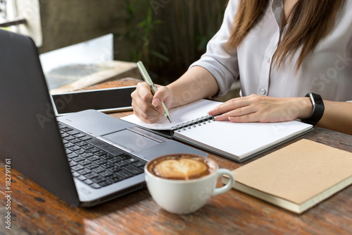 Business women using laptop and note some data on notepad, On table is laptop, smartphone and cup of coffee
