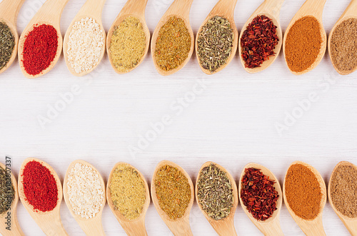 Decorative border of different spices in bamboo spoons on white wood background.