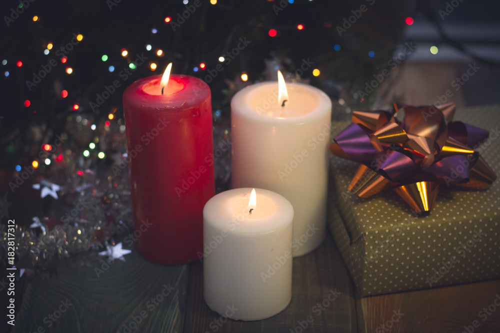 A festive still life with a red and two white candles of different size and a handcrafted gift box with a purple and orange shiny bow on a colored wooden surface. Dark Christmas background. Blurred A festive still life with a red and two white candles of different size and a handcrafted gift box with a purple and orange shiny bow on a colored wooden surface. Dark Christmas background. Blurred