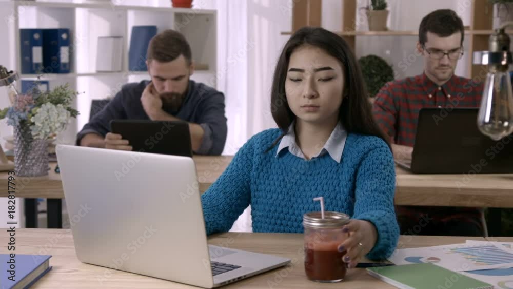 Attractive asian girl working with documents and laptop in open space office with coworkers using digital devices on background. Young female freelancer drinking healthy berry smoothie at workplace. Attractive asian girl working with documents and laptop in open space office with coworkers using digital devices on background. Young female freelancer drinking healthy berry smoothie at workplace.