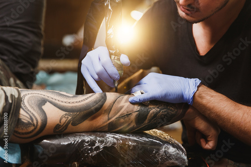 Close up of professional tattooer artist doing picture on hand of man by machine black ink from a jar. Tattoo art on body. Equipment for making tattoo art. Master makes tattooed in light studio. photo