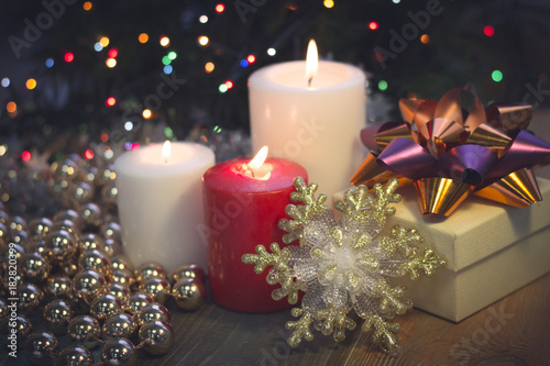 A festive still life with a red and two white candles of different size  a beige gift box with an orange and purple shiny bow  gold beads and toy snowflake. Dark Christmas background. Blurred bokeh