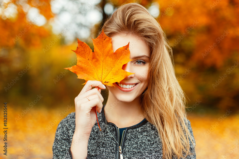Beautiful happy woman with a smile holds an autumn yellow leaf near the face - obrazy, fototapety, plakaty 